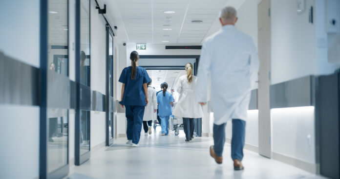 Hospital Hallway with Doctors, Nurses and Specialists in Hospital. Female and Male Physicians, Surgeons, Healthcare Officials Walk Together in Corridor with Their Back to Camera